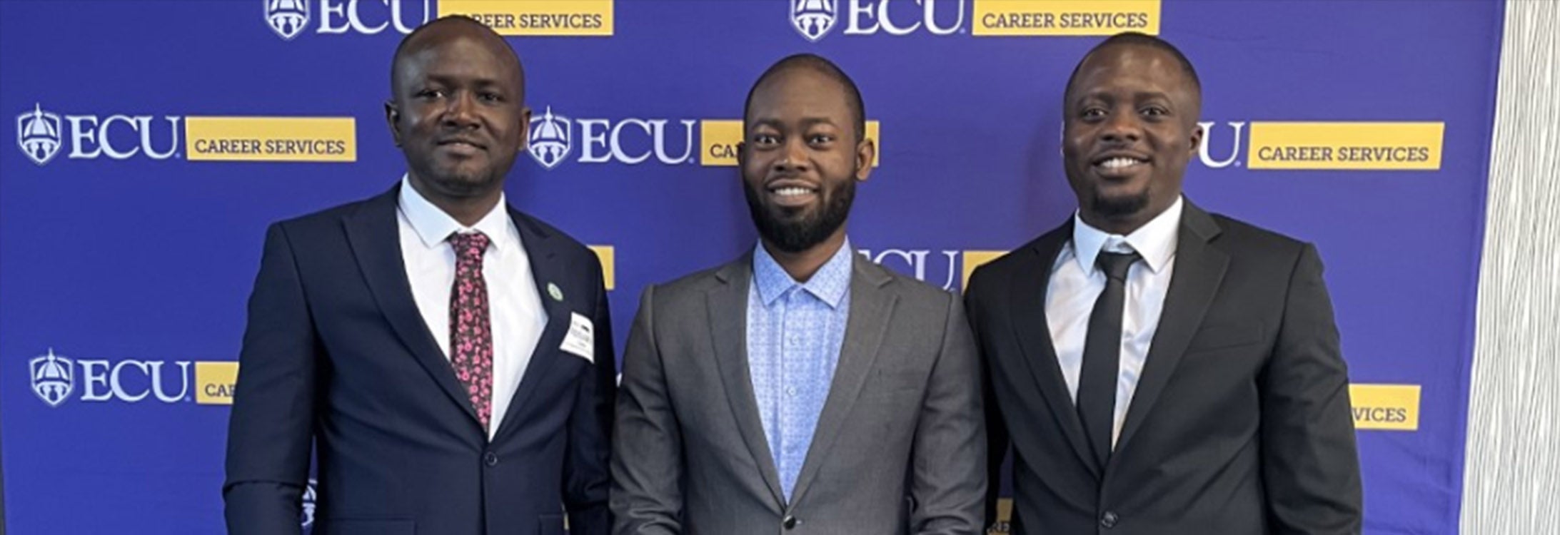 Three gentlemen smiling in front of ECU banner at Career Fair.